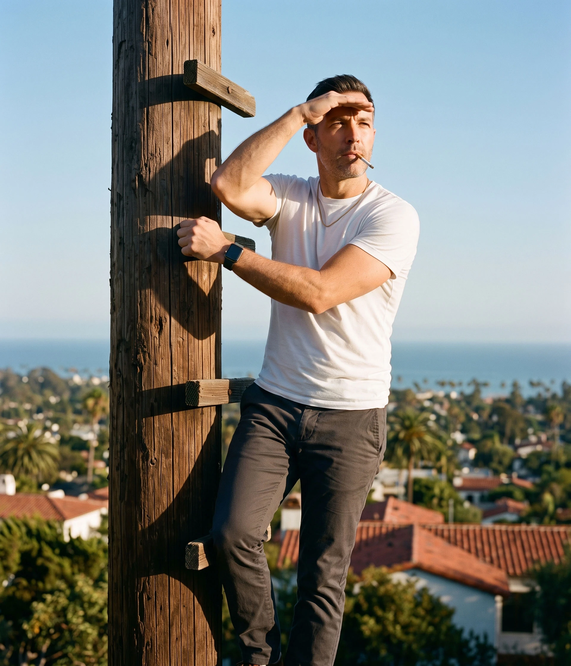 Man in a white t-shirt leaning on a wooden pole and shielding his eyes while looking out over a coastal town.